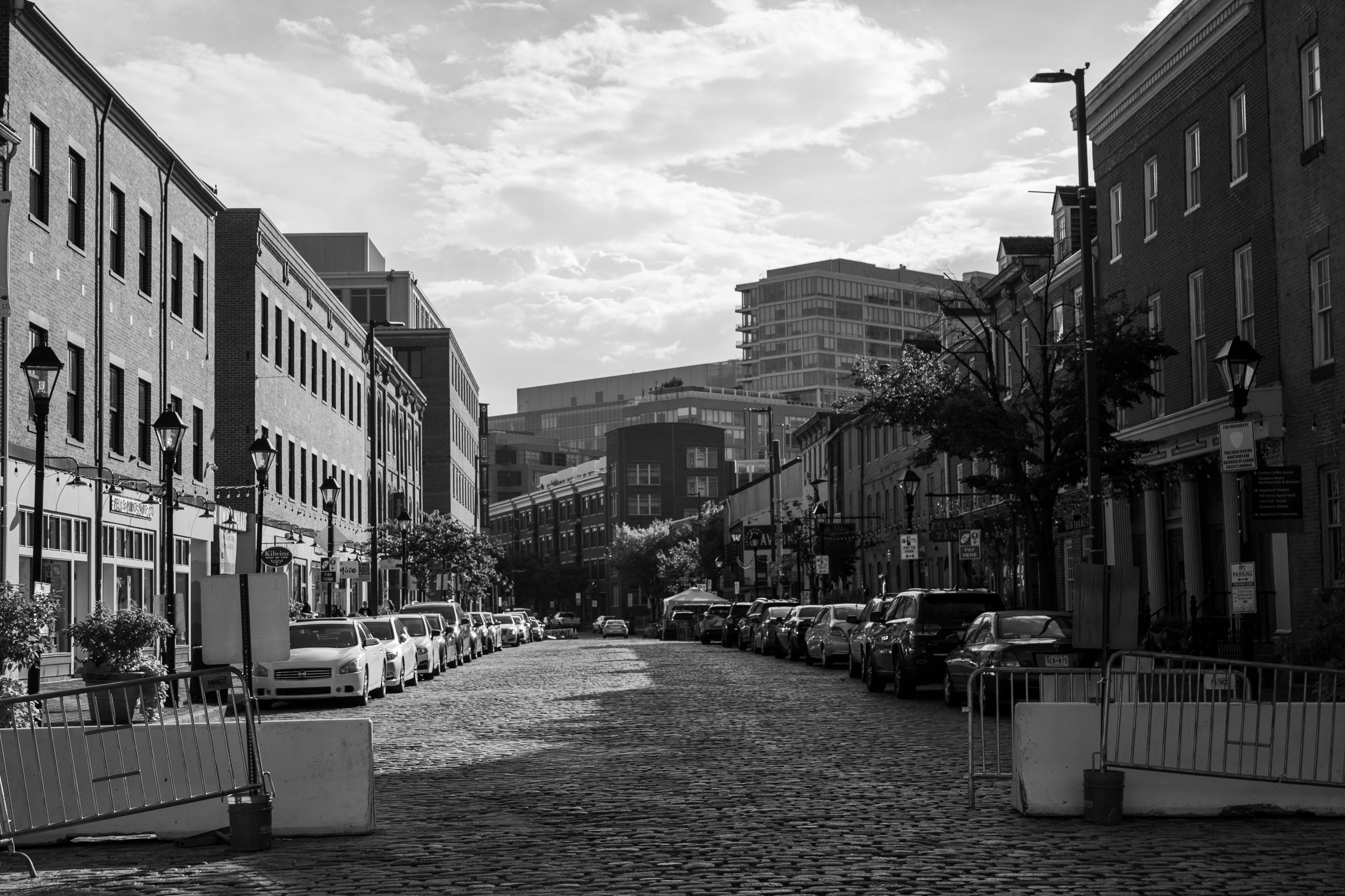 A black and white photo of a cobblestone city street lined with parked cars and brick buildings, barricades on both sides, and modern buildings visible in the background under a partly cloudy sky.